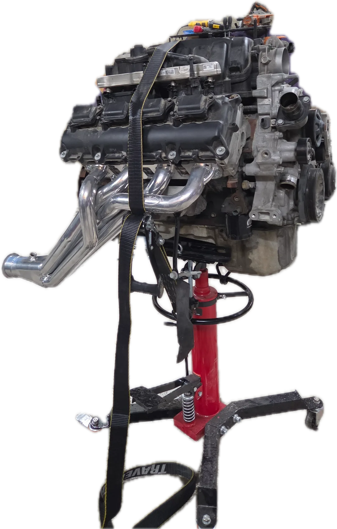An engine with polished long-tube headers, strapped to a red transmission jack in the MMKM shop