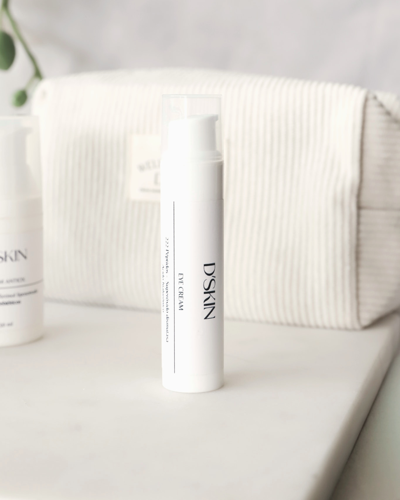 Frasco blanco de Eye Cream de D'SKIN sobre una repisa de mármol, con un neceser de lino claro al fondo