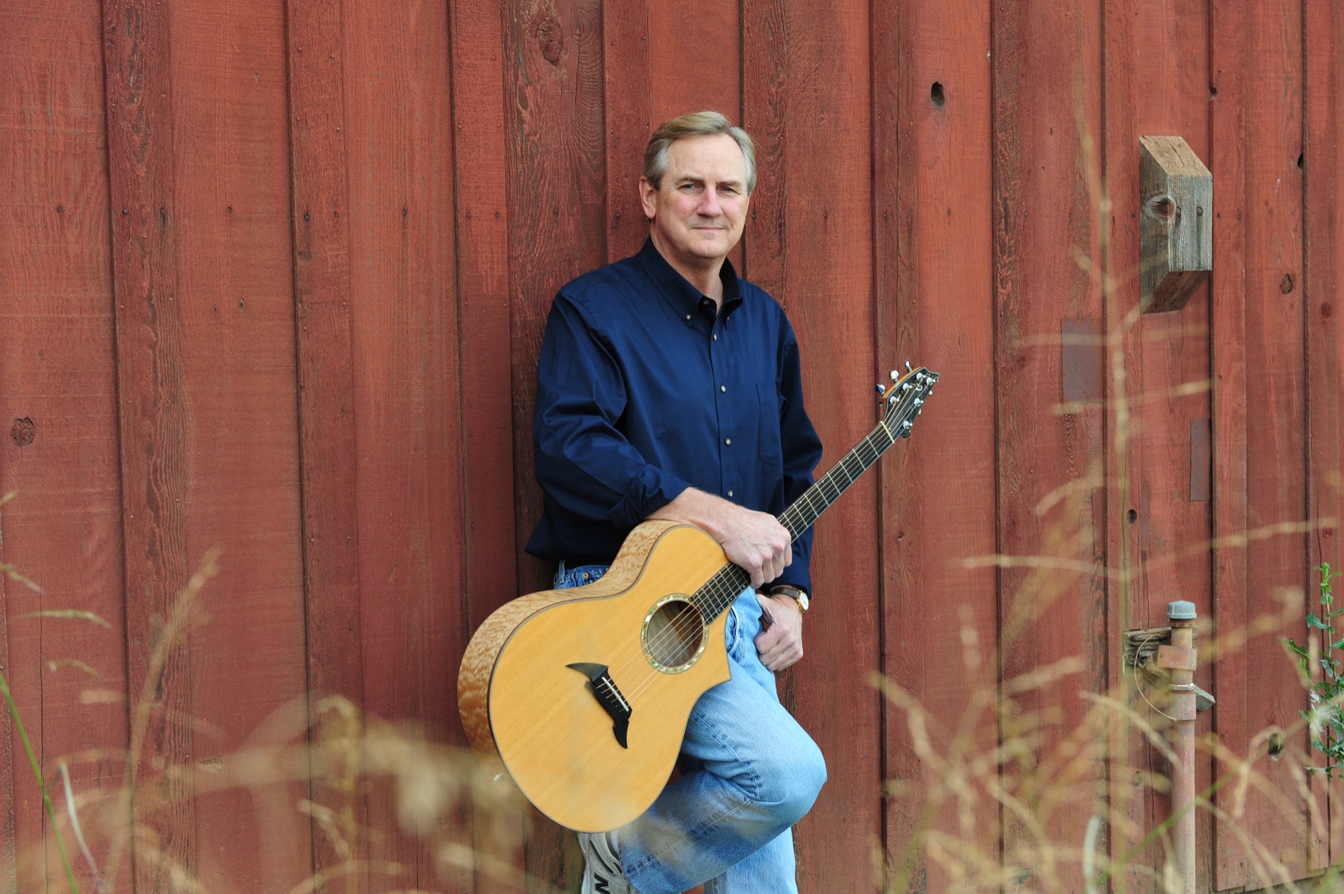 Kyle Freeman seated with his acoustic guitar in front of a red barn and tall grass
