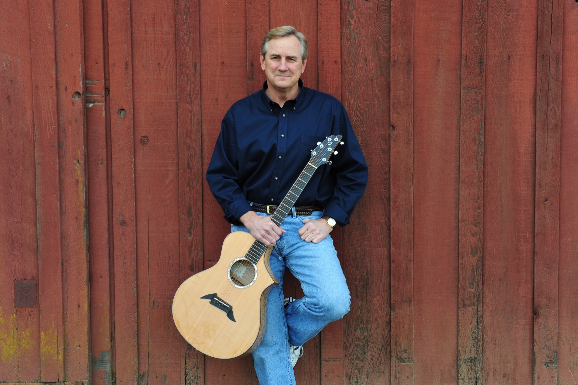 Kyle Freeman resting his acoustic guitar on his knee against a red barn wall