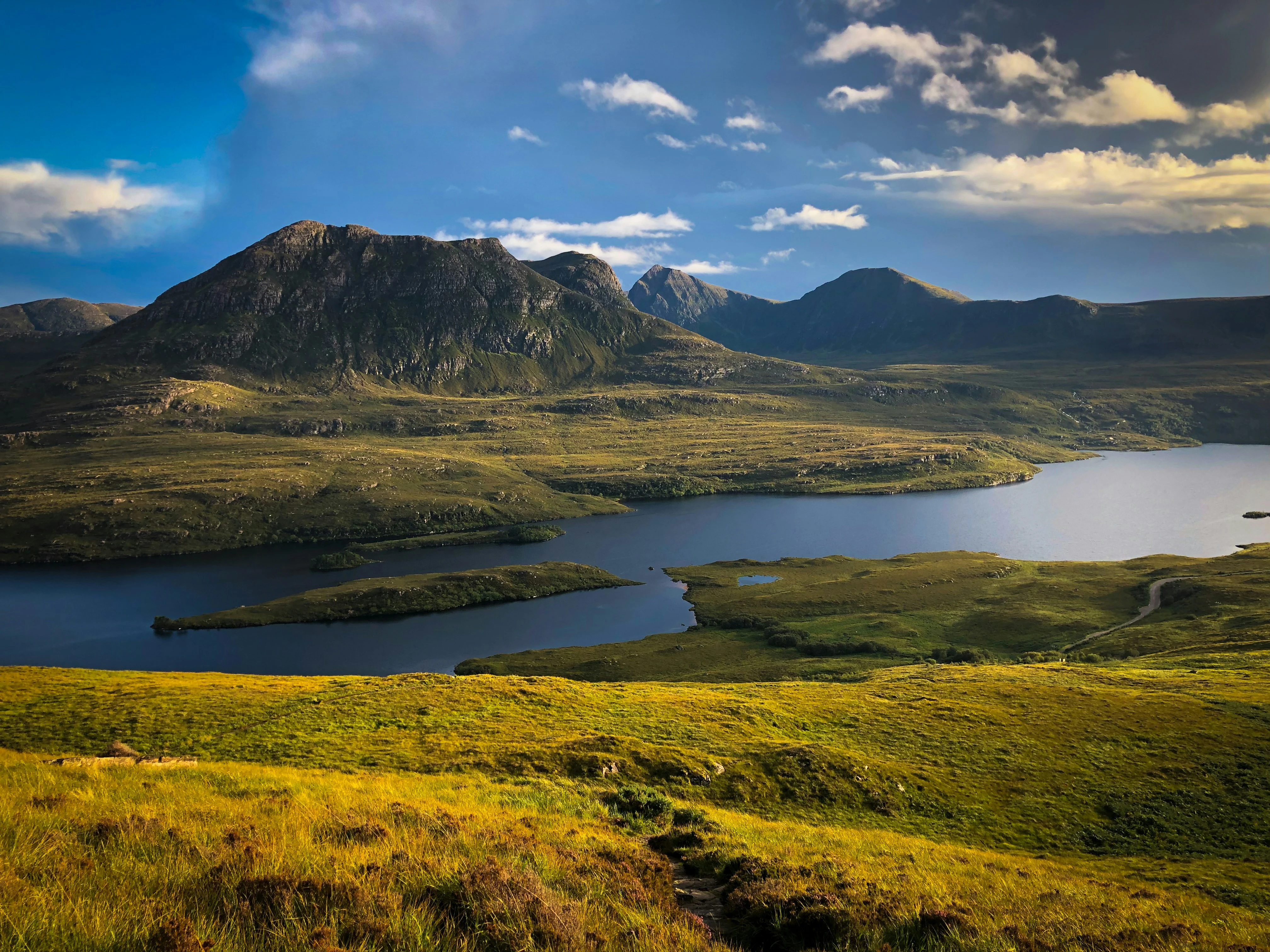 Mountain in the Scottish Highlands with a rocky peak and green valleys, overlooking a clear lake reflecting the bright sunlight under a sunny sky.