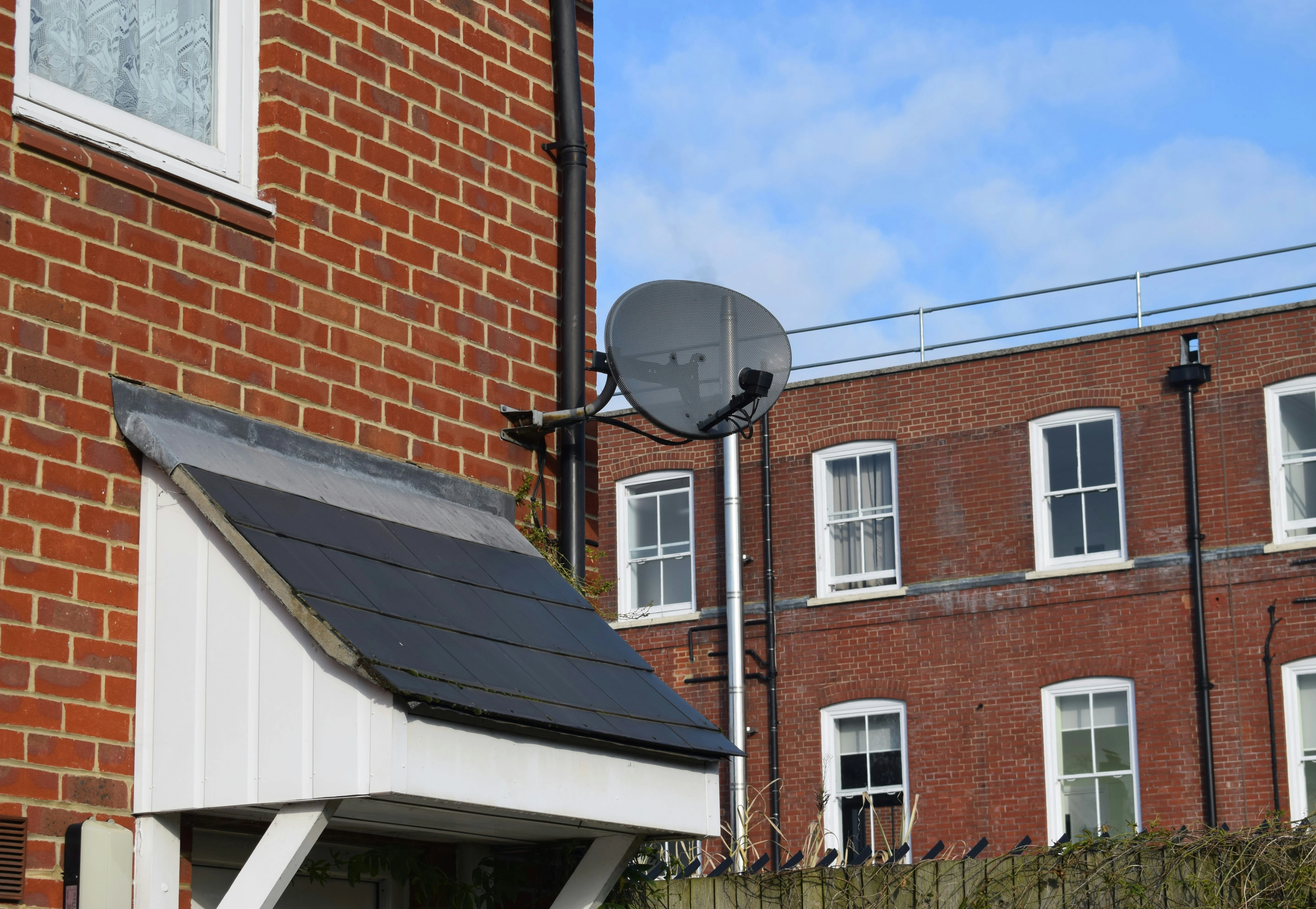 Close-up of a brick building with a small roofed entrance, featuring a black satellite dish mounted on the wall. In the background, another brick building with white-framed windows is visible under a blue sky with scattered clouds.