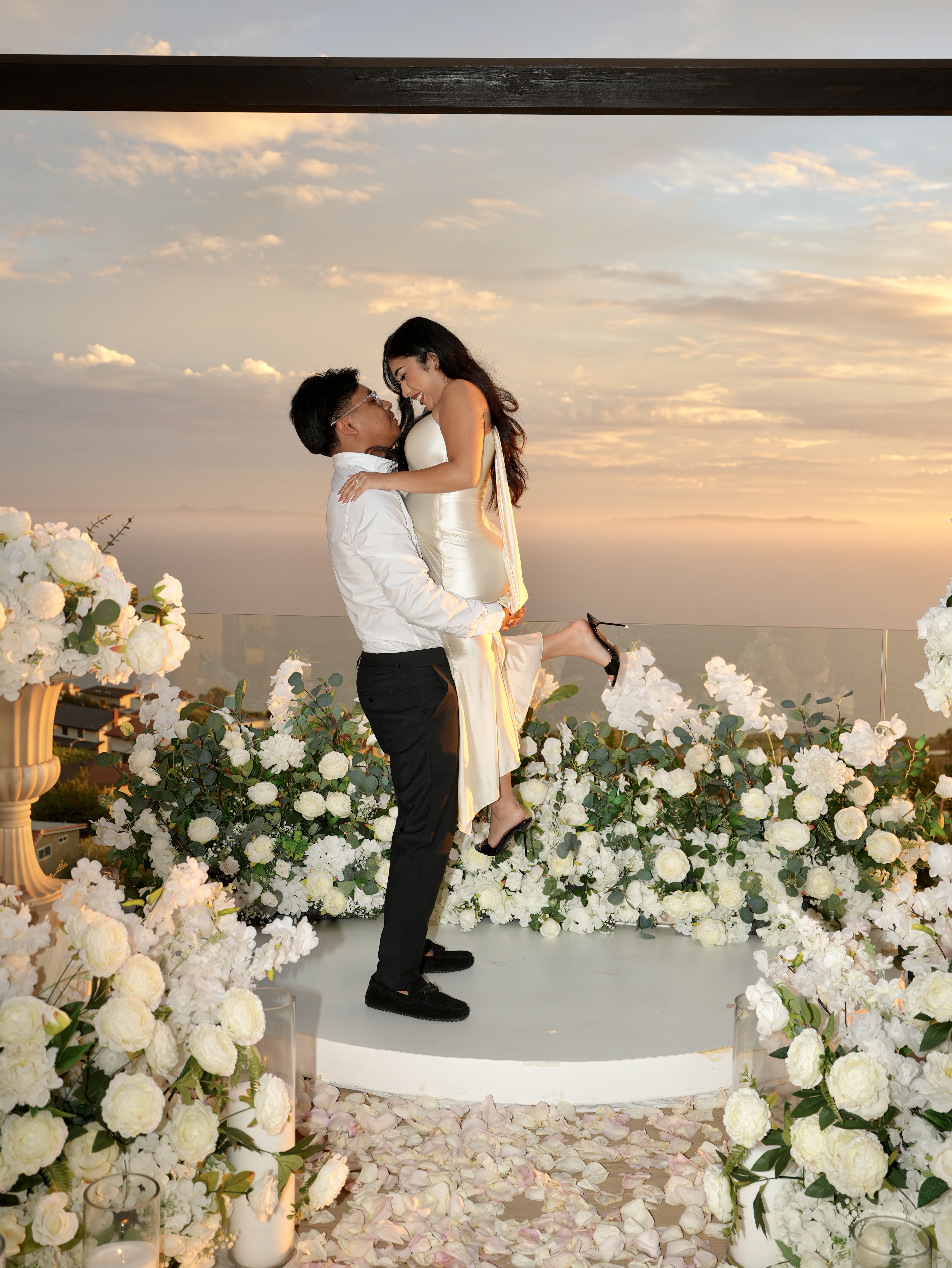 Romantic proposal surrounded by pink and white flowers, candles, and a scenic oceanfront backdrop.