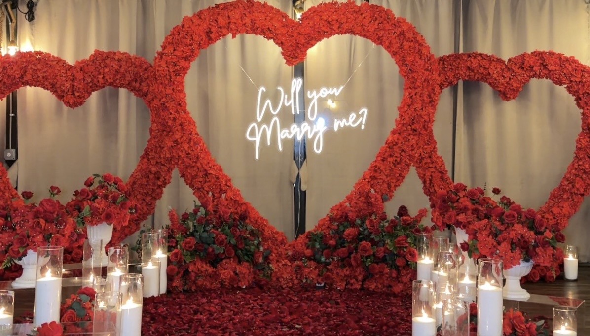A large blush-pink floral heart surrounded by roses, greenery, and glowing candles against a white draped backdrop.