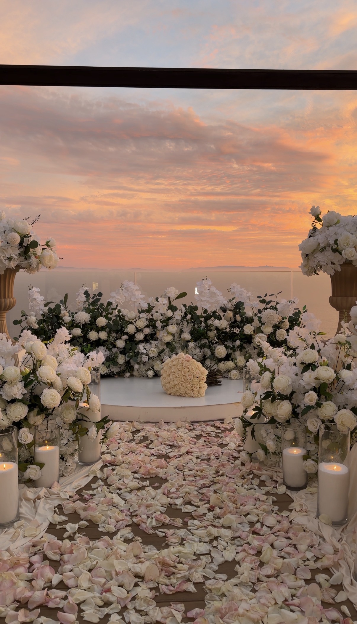 Elegant white floral arrangements overlooking the ocean, with a petal-lined aisle, candles, and a romantic sunset setting.