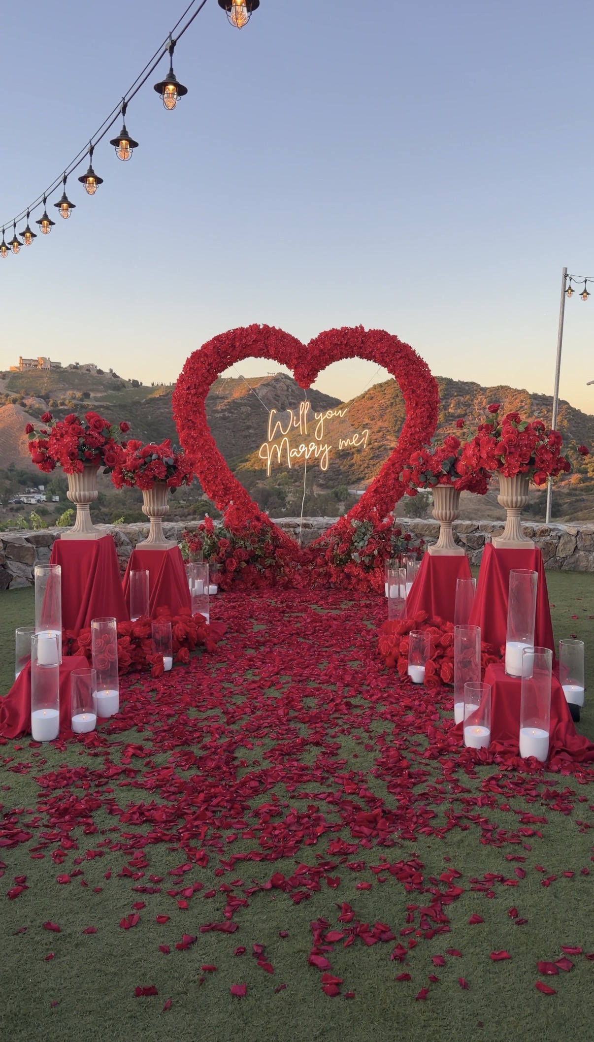 A vibrant red floral heart backdrop with a “Will You Marry Me?” sign, red roses, candles, and elegant outdoor greenery overlooking the hills.