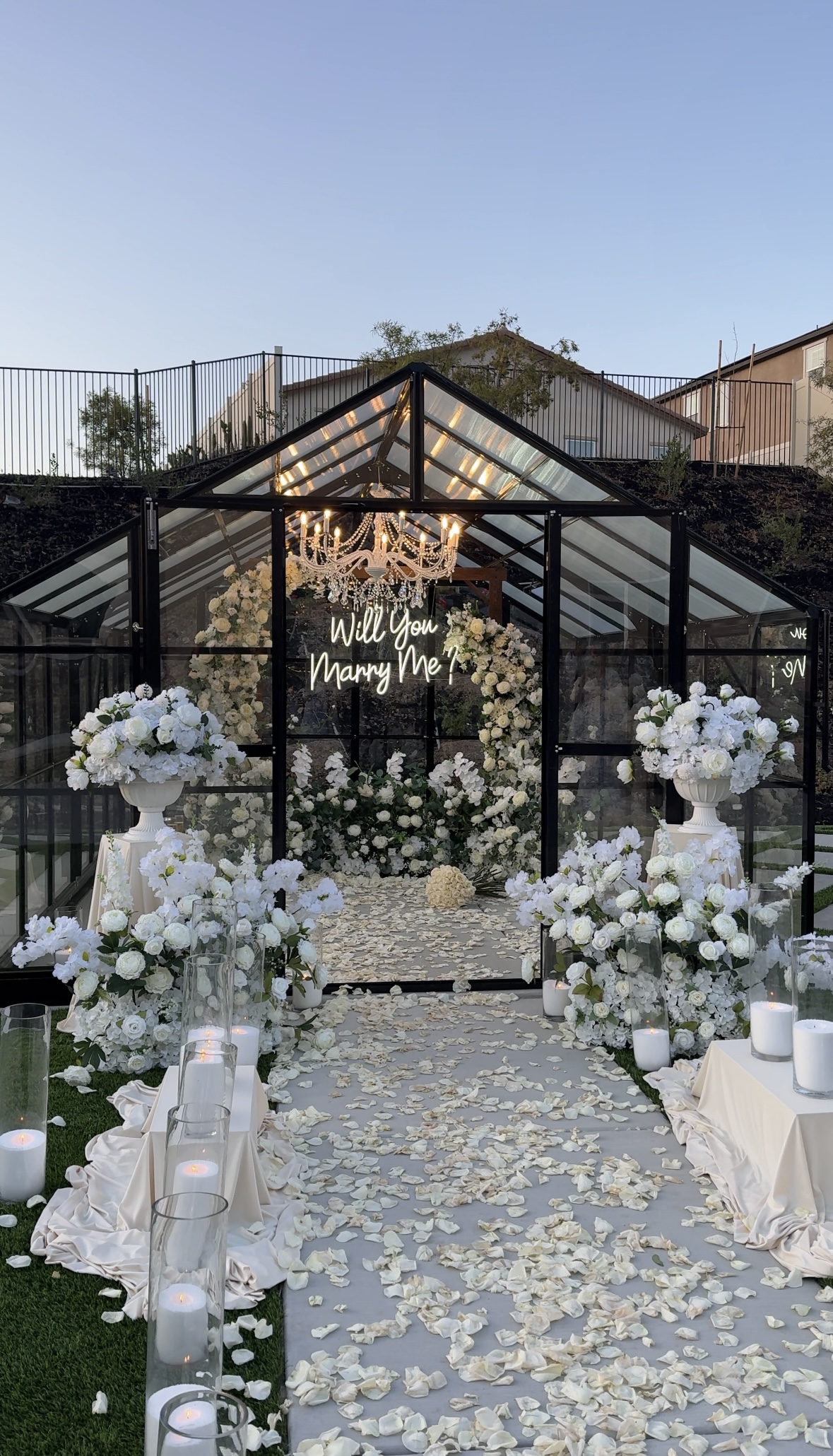 A glass greenhouse decorated with white florals, candles, a crystal chandelier, rose petals, and a glowing “Will You Marry Me?” sign.
