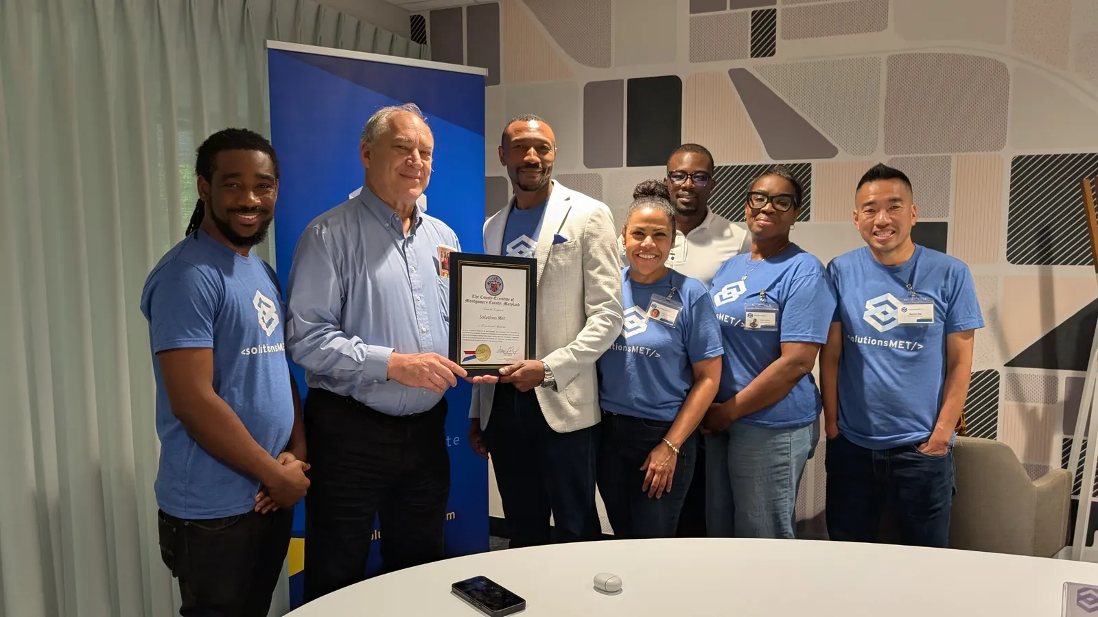 Group posing with a framed certificate in front of a SolutionsMET banner.