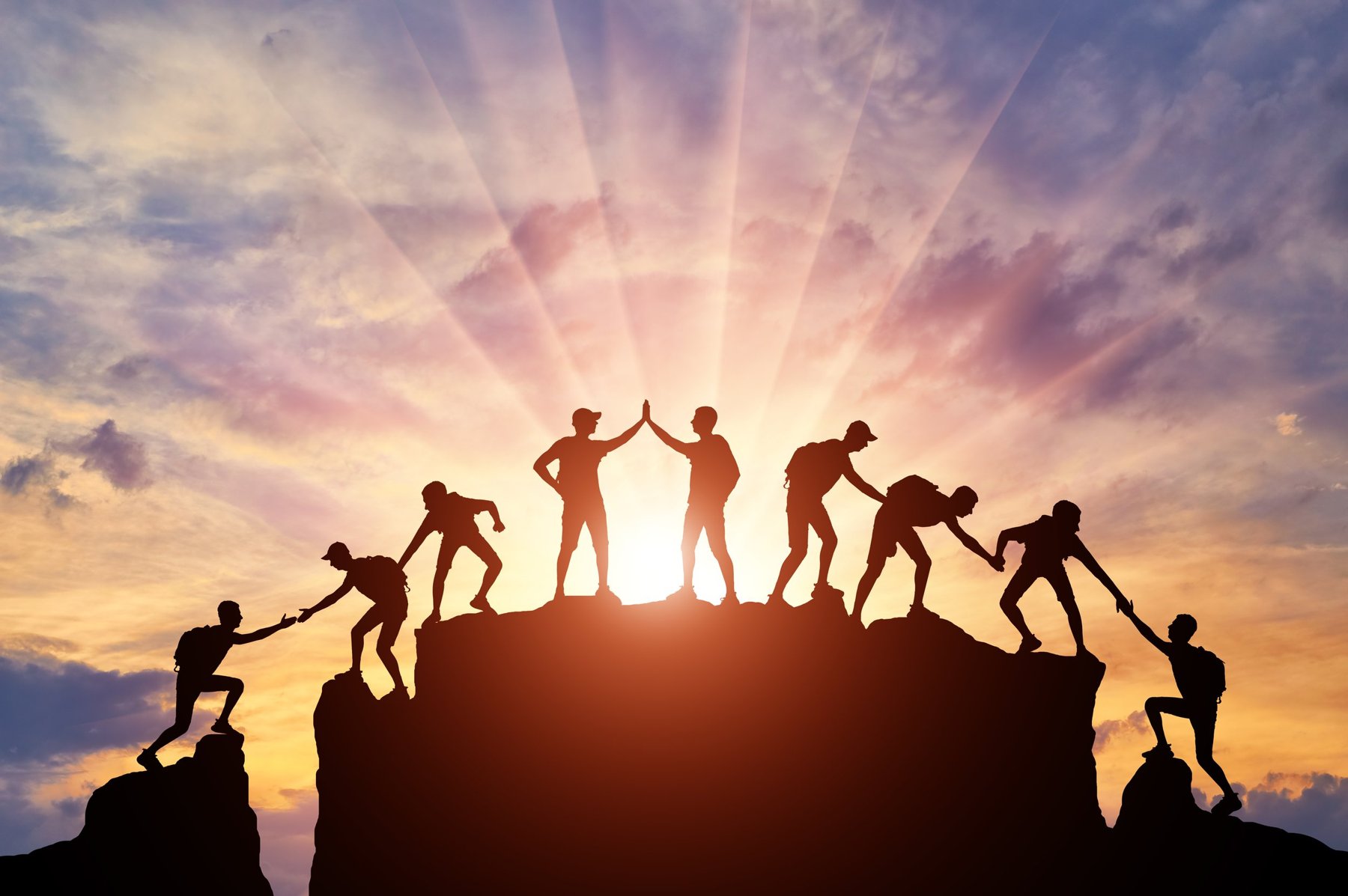 Silhouettes of a team helping each other up a mountain at sunset