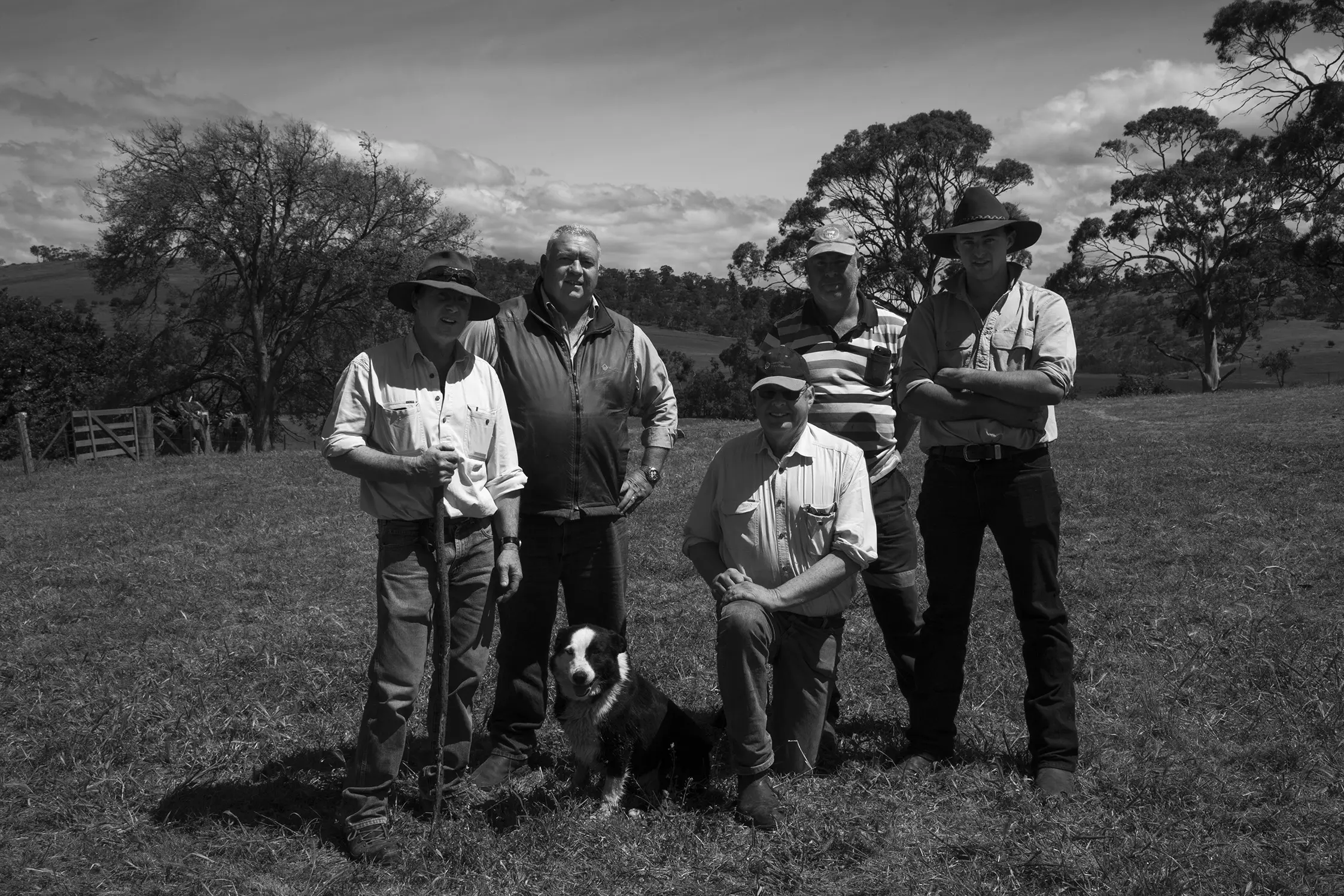 A group of farmers standing together in a field, posing for a photo with a sheepdog in front of them.