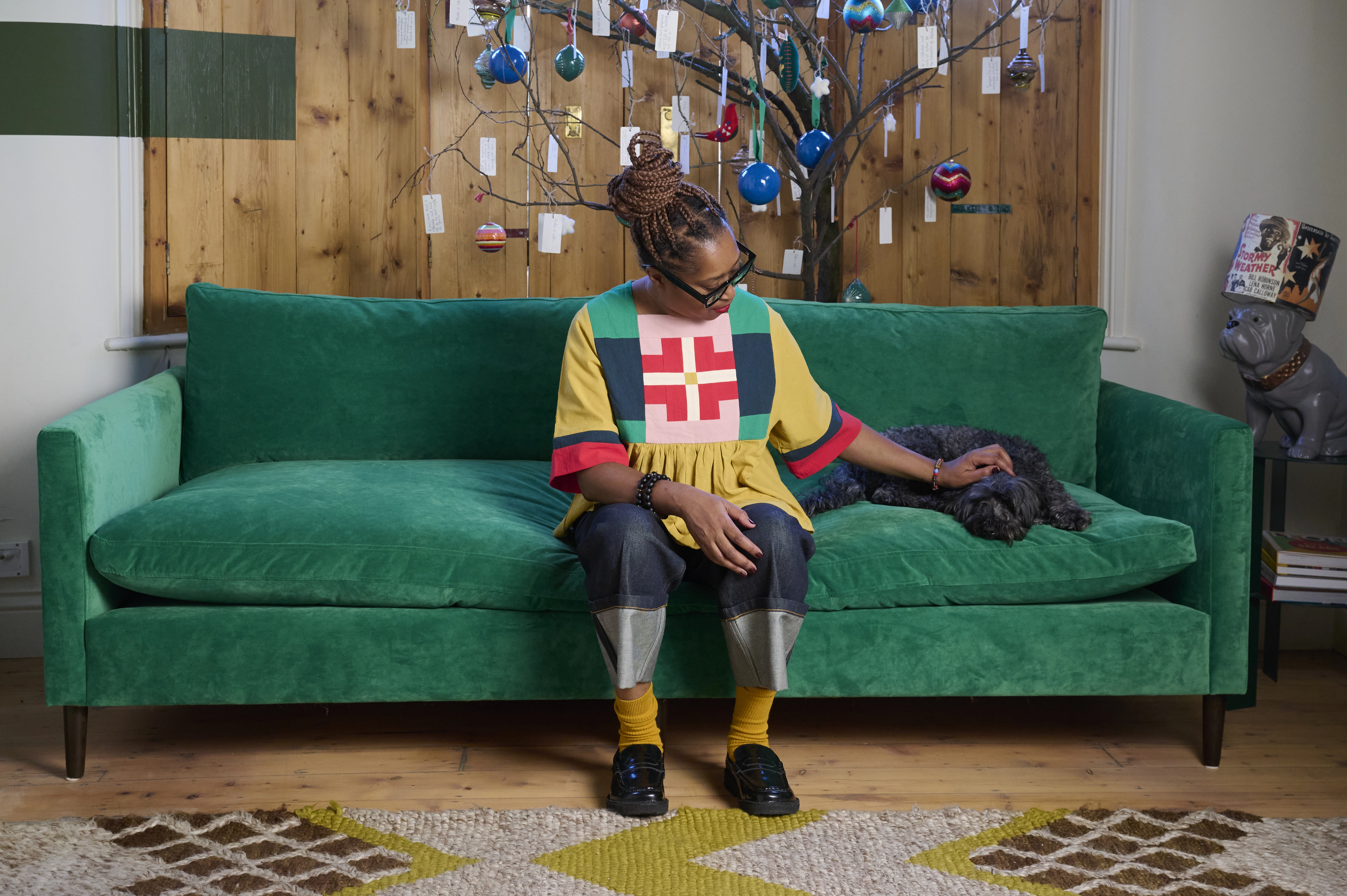 A person sits on a green sofa, gently petting a dog in a cosy living room.