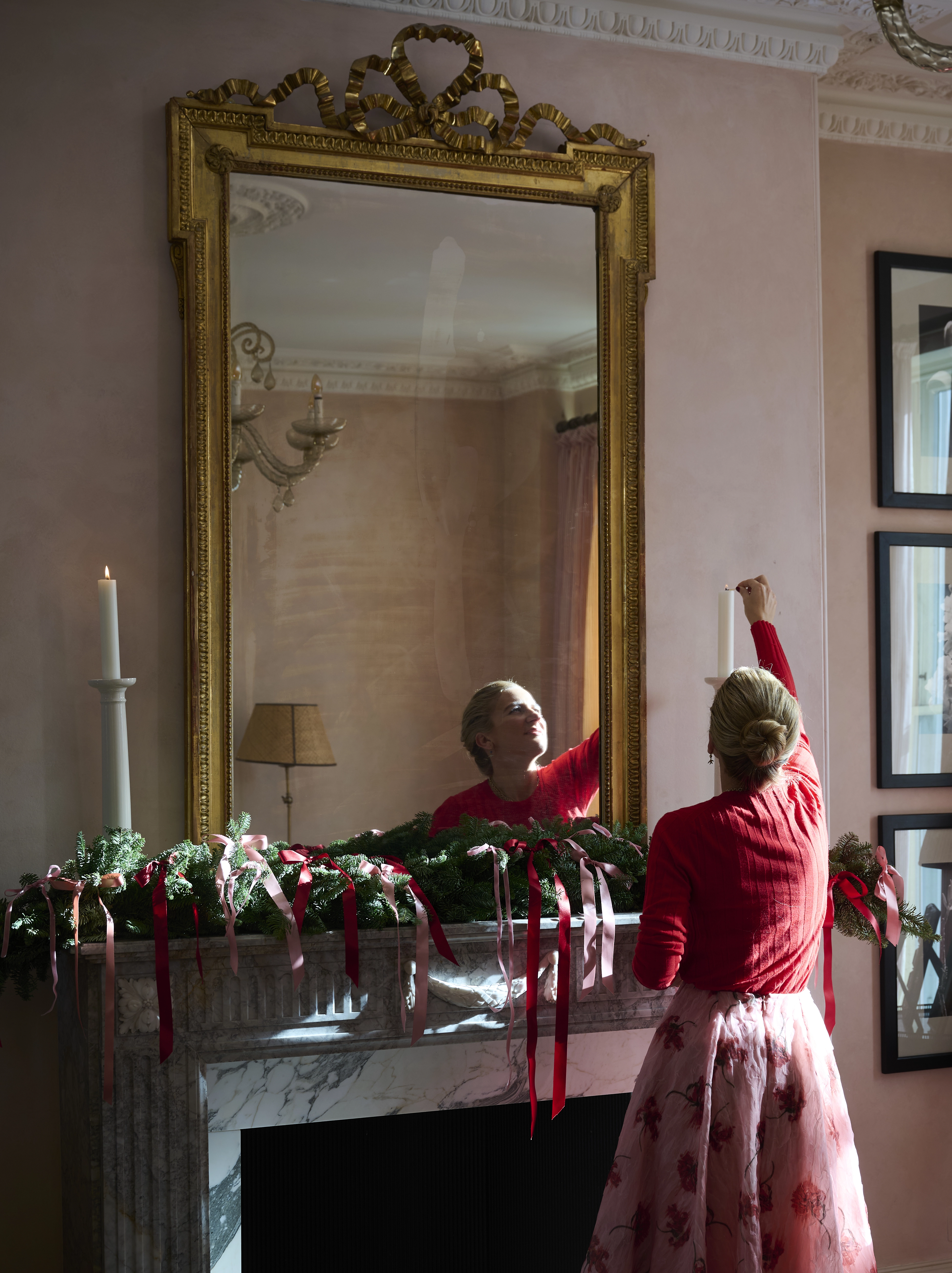 A woman lights a candle on a decorated fireplace beneath a large mirror.