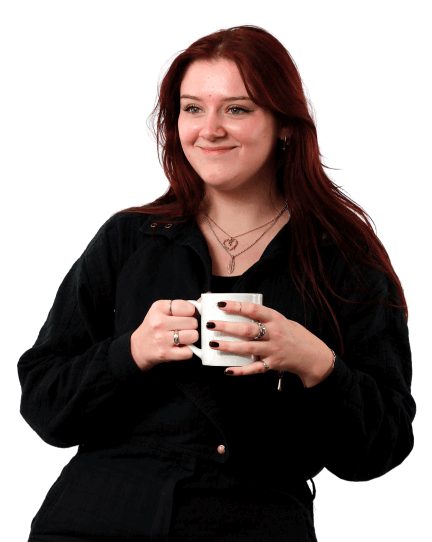 Katie is wearing black trousers and a black jacket as she sits casually on a wooden stool holding a coffee mug. She’s looking off-camera and smiling.