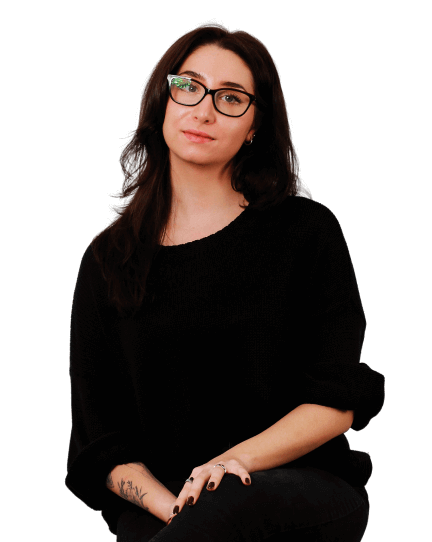 Angie is wearing black jeans, black glasses and a black cardigan. She’s sitting on a stool with her head tilted to one side. 