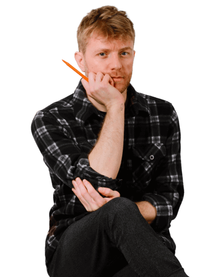 Jim is wearing a plaid shirt and black trousers as he sits on a wooden stool with his legs crossed. He holds an orange pen and looks directly into the camera, attempting to look scholarly.