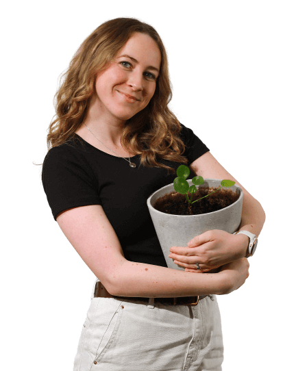 Fiona is wearing beige trousers and a black t-shirt as she lovingly hugs a pot plant and looks directly into the camera smiling.