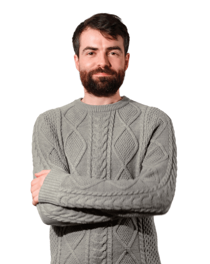 Greg is wearing a grey jumper and black trousers. He stands with his arms crossed, smiling slightly and looking directly into the camera. 