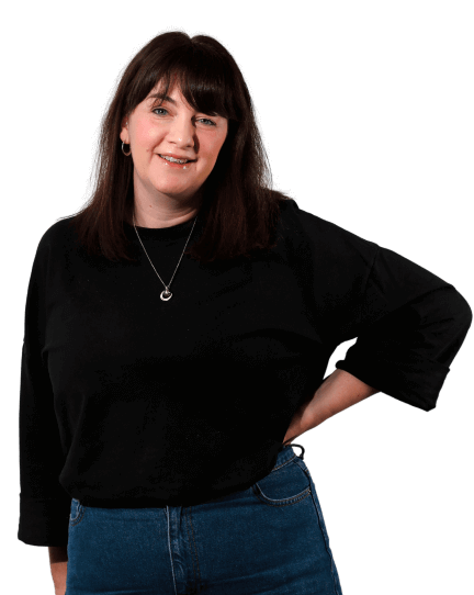 Siobhan is wearing denim jeans and a black cardigan as she leans on a wooden stool with one hand and looks directly into the camera. 