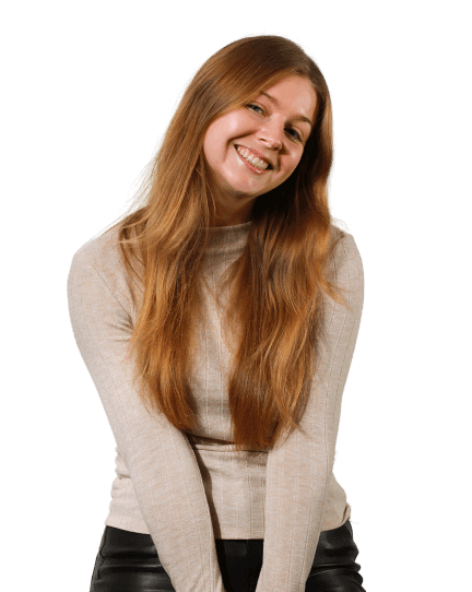 Jess is wearing black trousers and a beige cardigan as she sits on a wooden stool and looks directly into the camera with a big smile, her head slightly tilted.