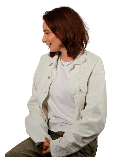 Grace is wearing olive green jeans with a white shirt and t-shirt as she sits on a stool and looks off-camera at the floor laughing. 