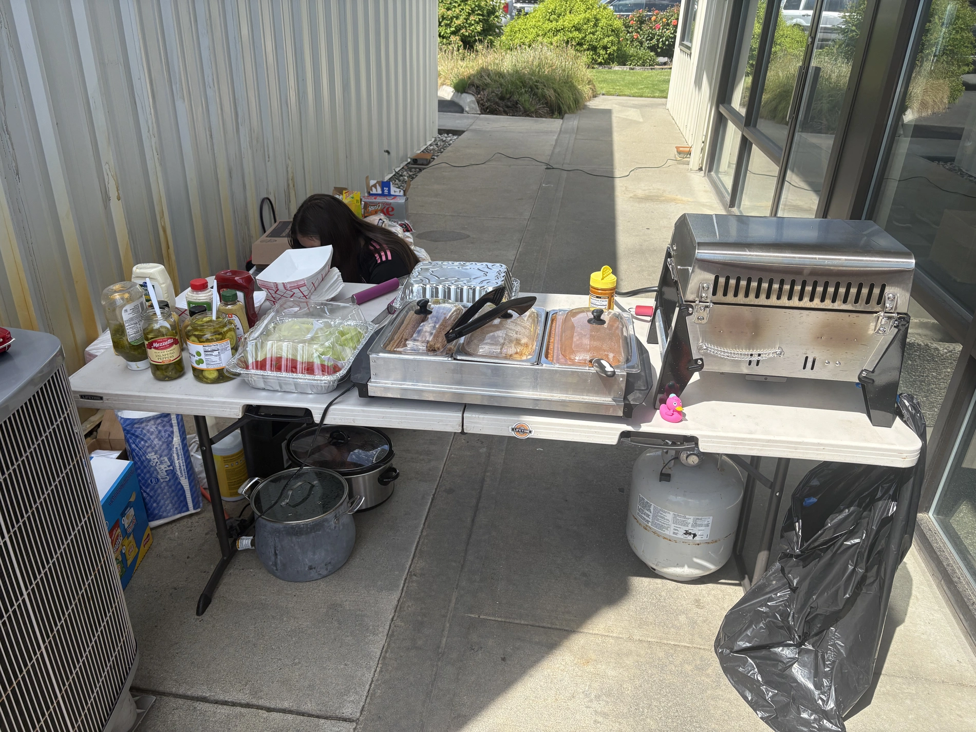 An outdoor food service station with a grill, slow cookers, and various condiments prepared for guests at a community event.