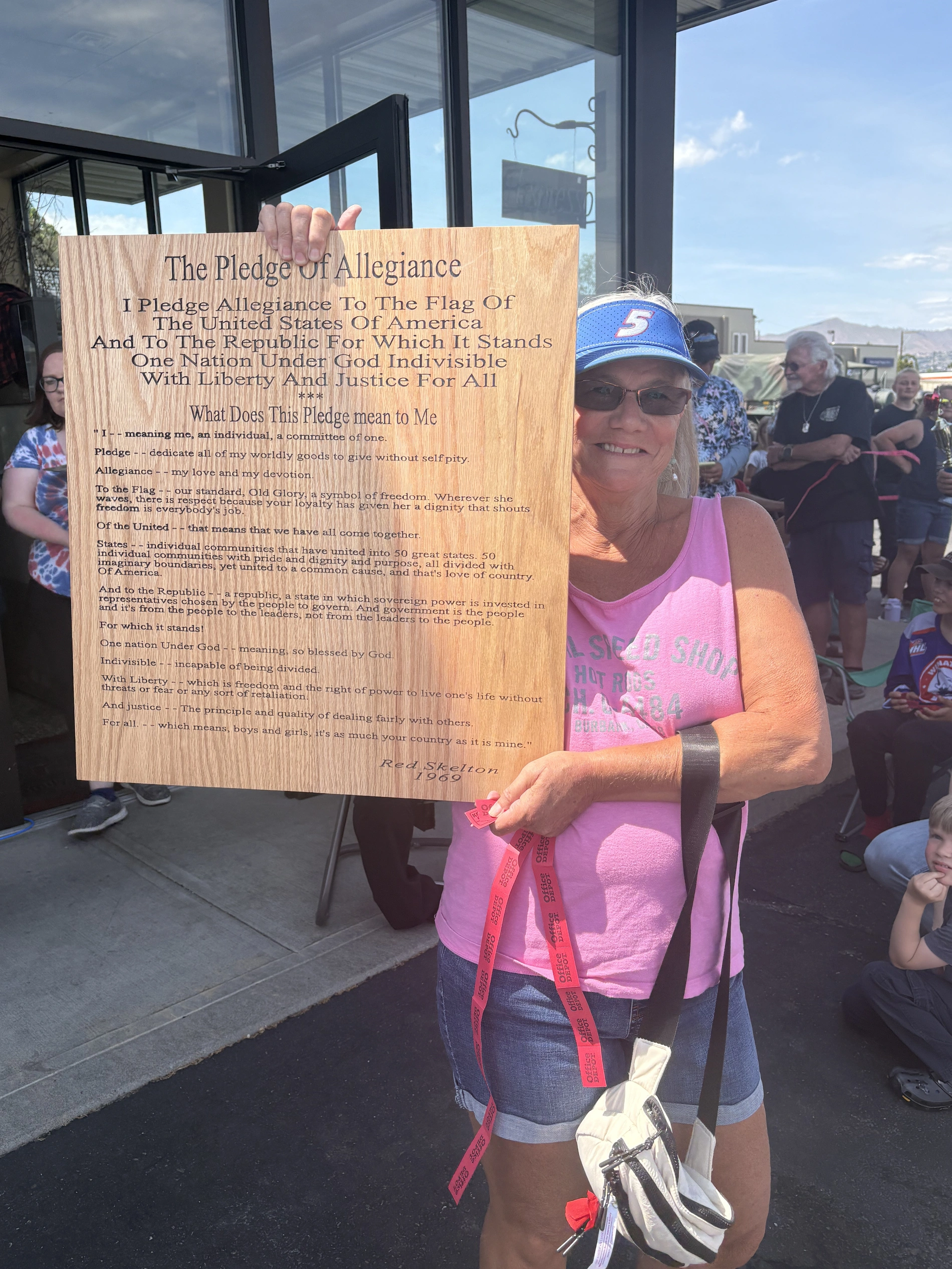 A woman smiling and holding a large wooden plaque engraved with the Pledge of Allegiance and Red Skelton’s 1969 commentary on its meaning.
